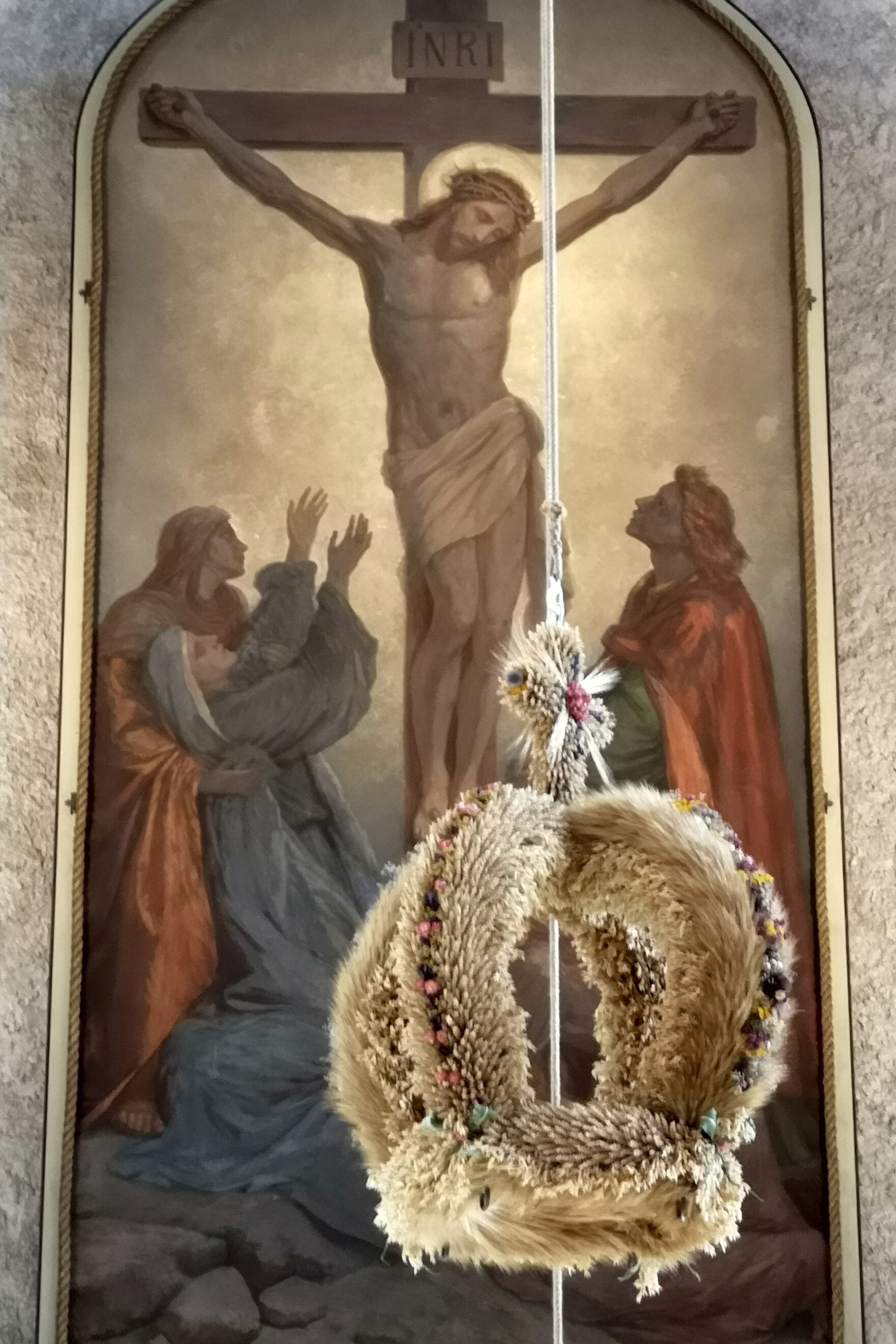 Erntekrone aus Getreide und Blumen mit Kreuz aus Getreide als Abschluss, schwebend vor Altarbild mit Kreuzigungsdarstellung