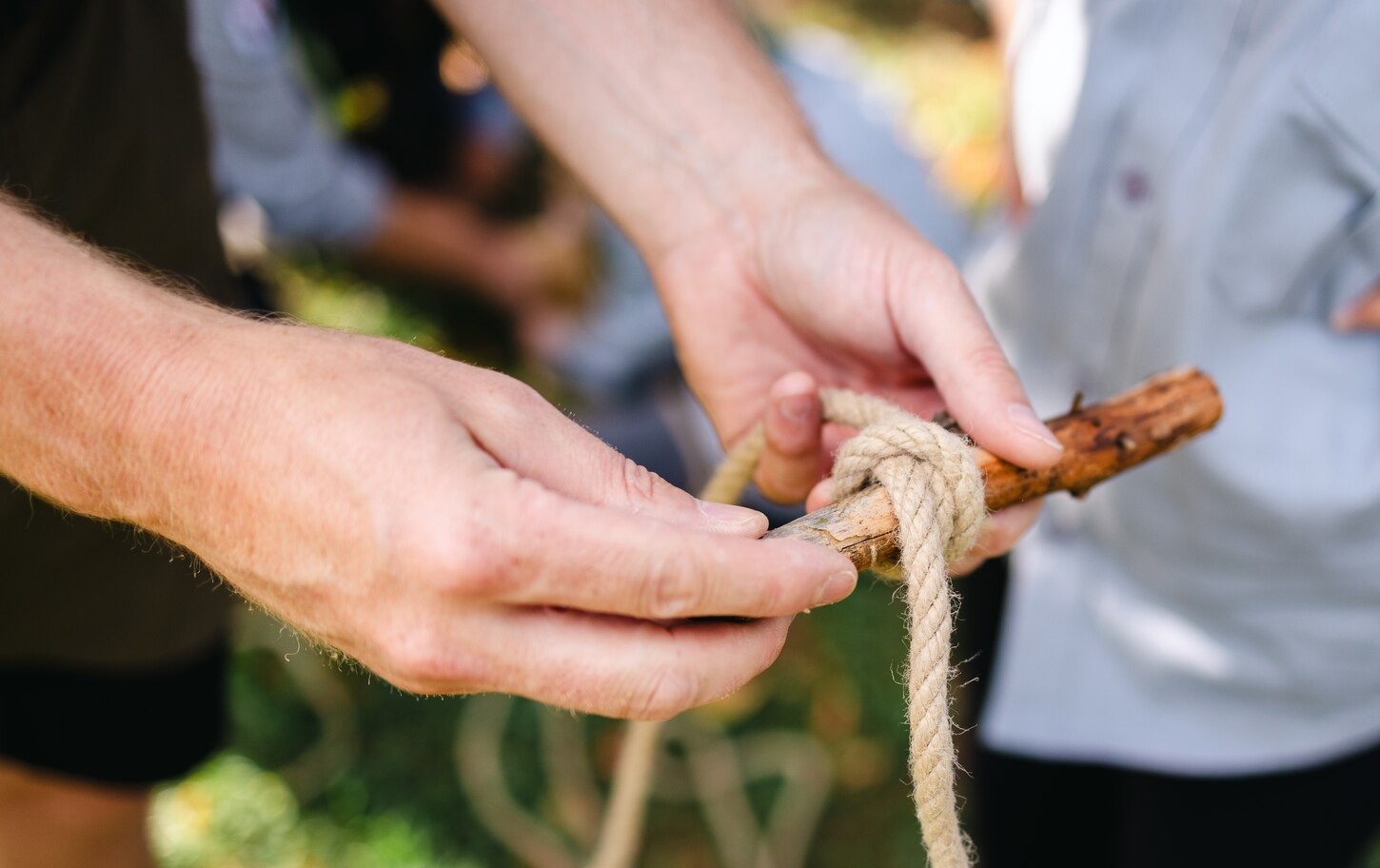 Eine Person zeigt einem Pfadfinderkind in Mülsen St. Micheln, wie ein Knoten gebunden wird. Detailaufnahme der Hände, die ein Seil um einen Stock wickeln.