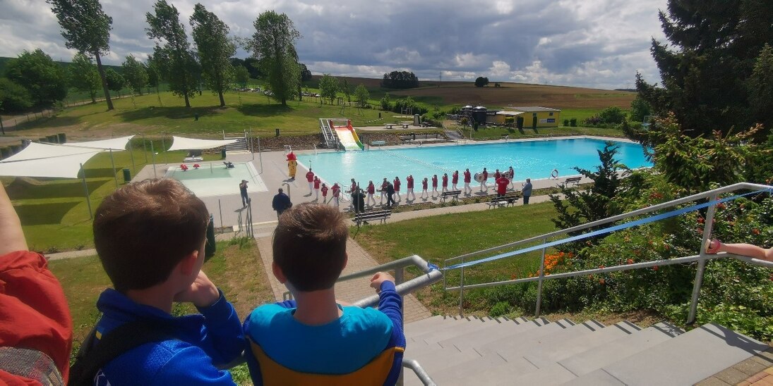Im Vordergrund zwei Kinder, die auf das Freibad schauen. im Hintergrund die Freifläche des Bades sowie ein Schwimmbecken.
