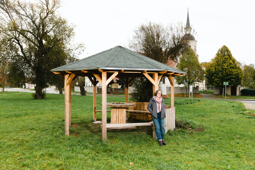 Frau steht vor einem Holzpavillion, im Hintergrund ist ein Kirchturm zu sehen