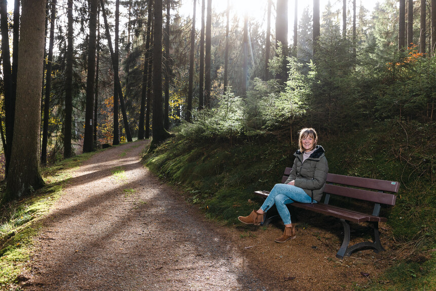 Eine Frau sitzt auf einer Holzbank an einem Waldweg. Sonne scheint durch die Bäume und es gibt ein Zitat der Vorsitzenden des Fremdenverkehrsvereins Bad Brambach e.V.