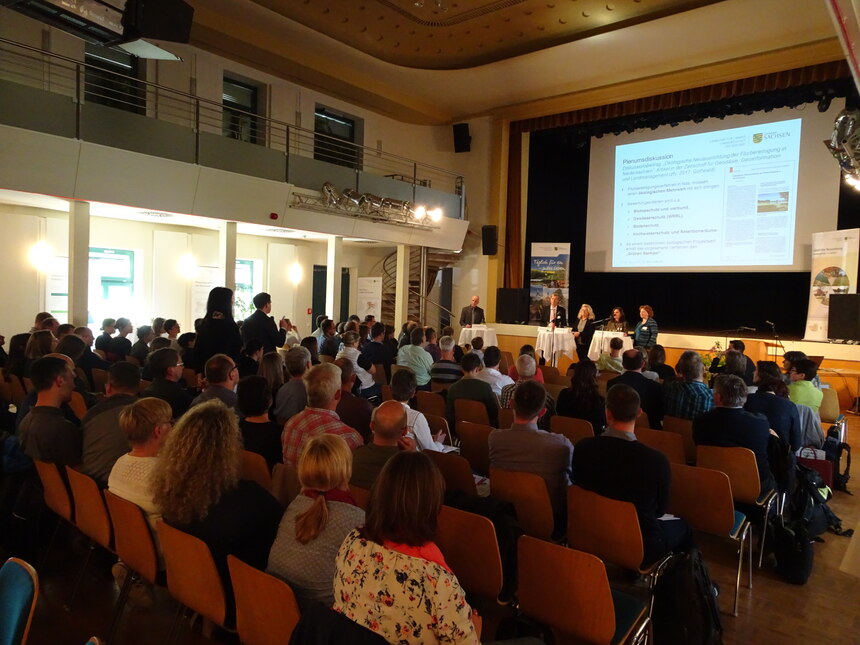 Blick von oben in den abgedunkelten Veranstaltungssaal des Bürgerhauses Geithain über die Köpfe der Teilnehmer hinweg Richtung Podium und die Leinwand