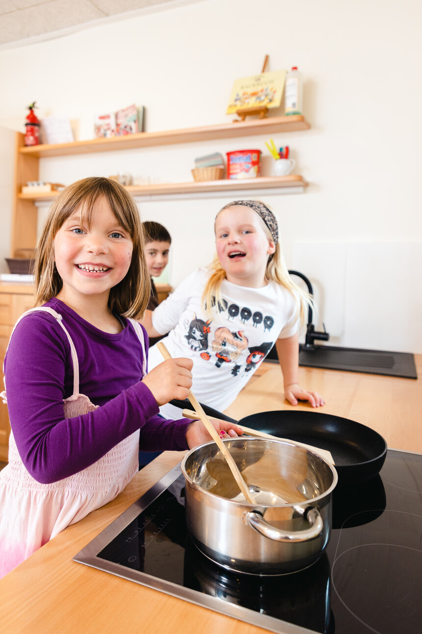 Kinder schauen fröhlich in die Kamera. Ein Kind rührt mit einem Holzlöffel im Kochtop