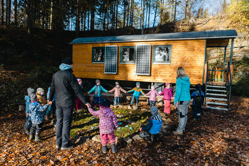 Zwei Erzieher stehen mit mehrern Kindern im Kreis im Freien vor dem Bauwagen und halten sich an den Händen.