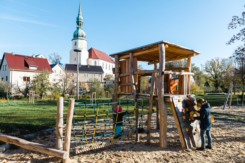 Frau und kleines Kind spielen auf dem Spielplatz. Im Hintergrund steht eine Kirche