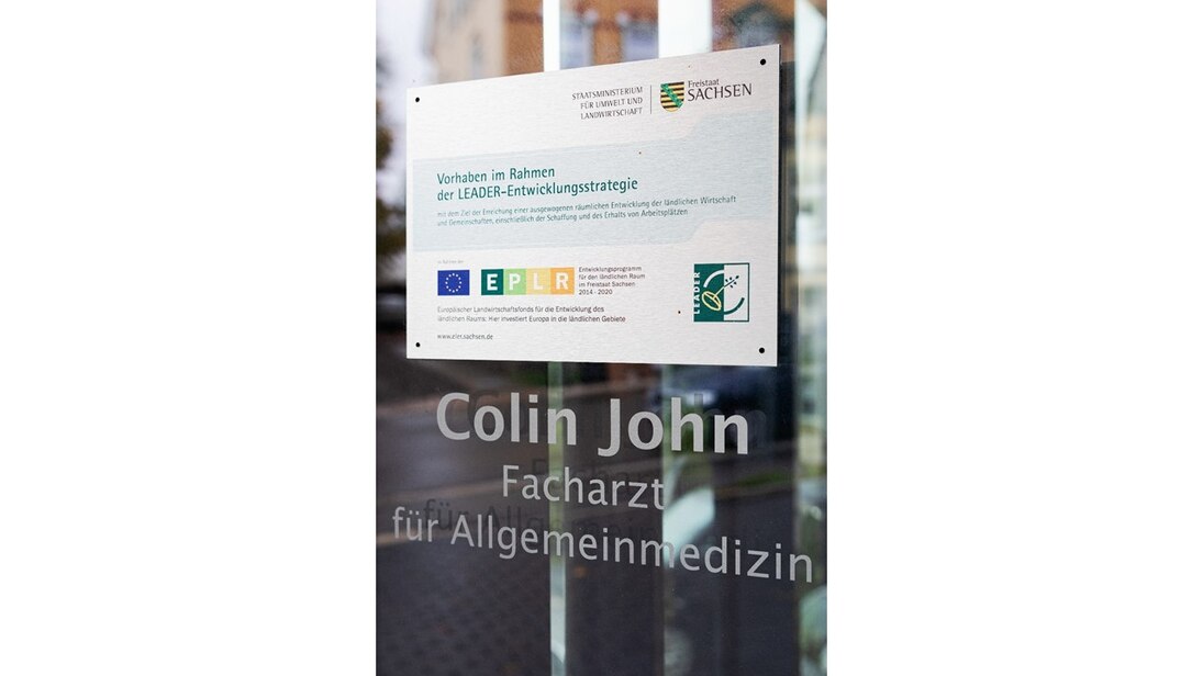 Glastür mit der Aufschrift Collin John Facharzt für Allgemeinmedizin und einem Förderhinweisschild Vorhaben im Rahmen der LEADER-Entwicklungsstrategie.