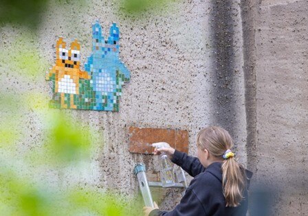 An einer Hauswand befindet sich ein gestaltetes Mosaikbild mit zwei Figuren. Im Vordergrund steht ein Mädchen mit einer Leiter.