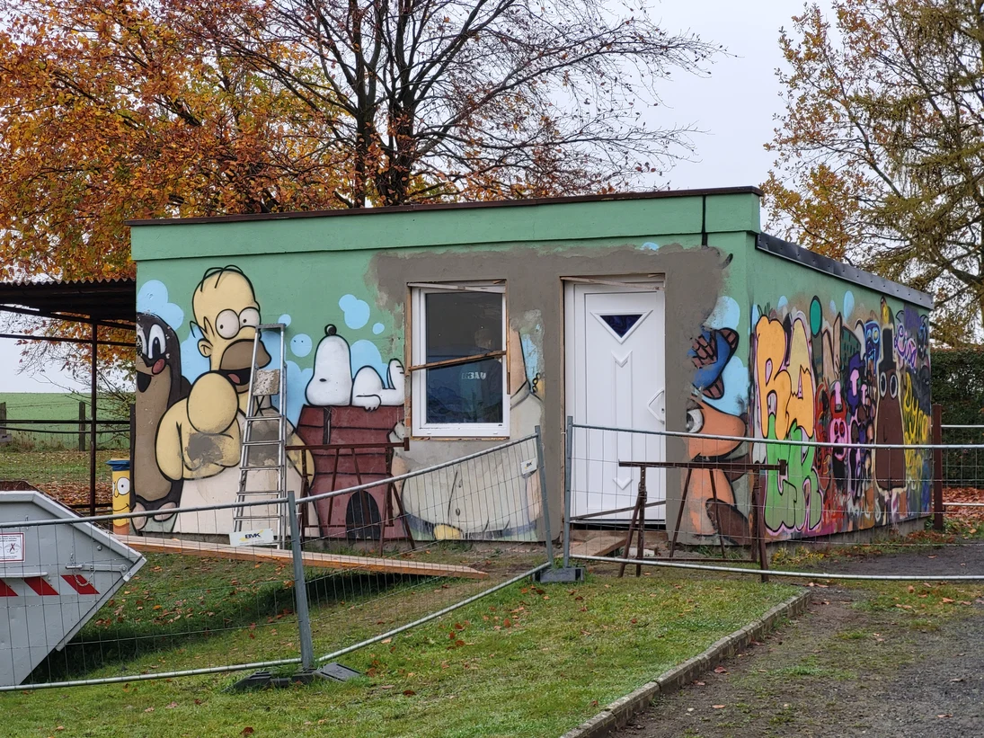Blick auf die Schulhütte. Die Fassade ist mit Graffiti gestaltet. Um die Hütte ist ein Bauzaun aufgebaut. Vorn links steht ein Container