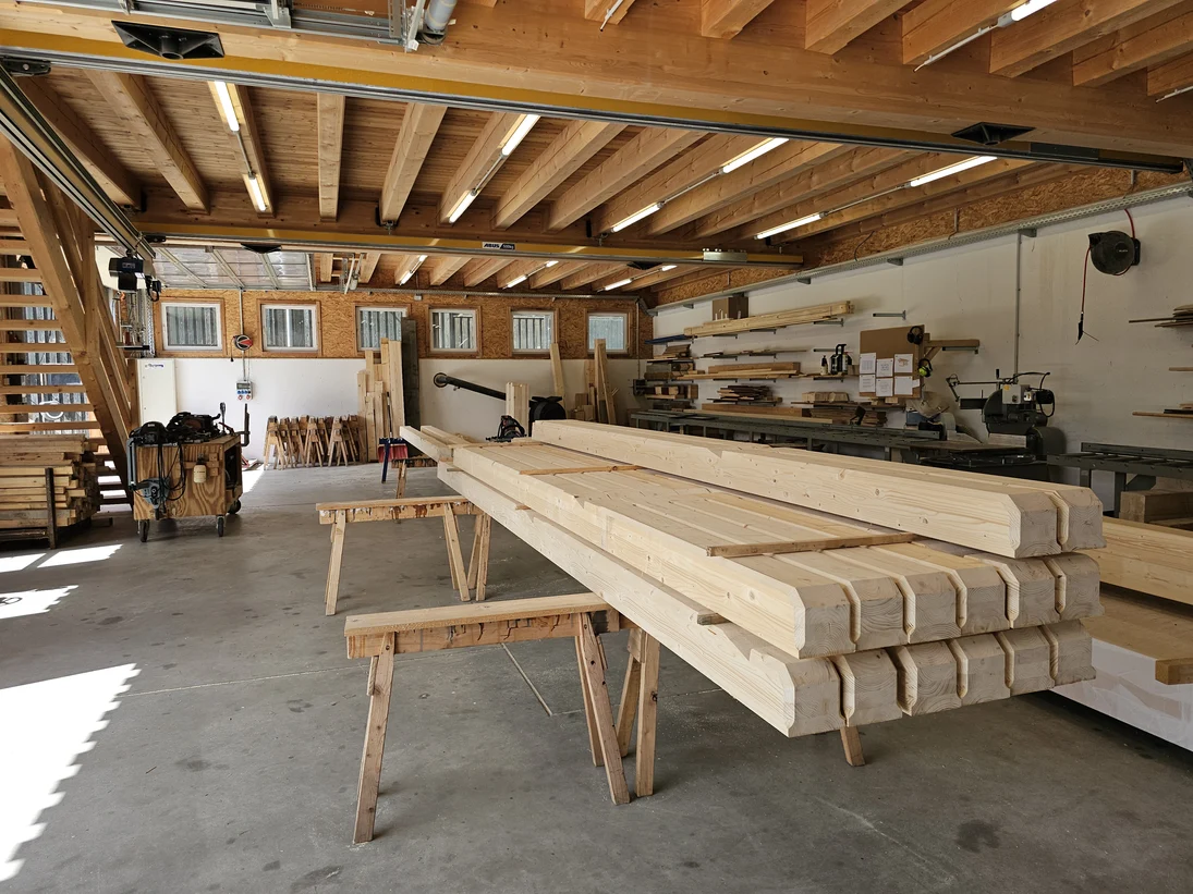 In der Halle lagern große Bauteile aus Holz. An den Wänden und der Decke befinden der Kran und Werkzeuge