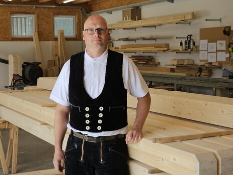 Firmeninhaber Martin Ritter steht in typischer Zimmermannskleidung vor einem großen Stabel aus bearbeitetem Holz