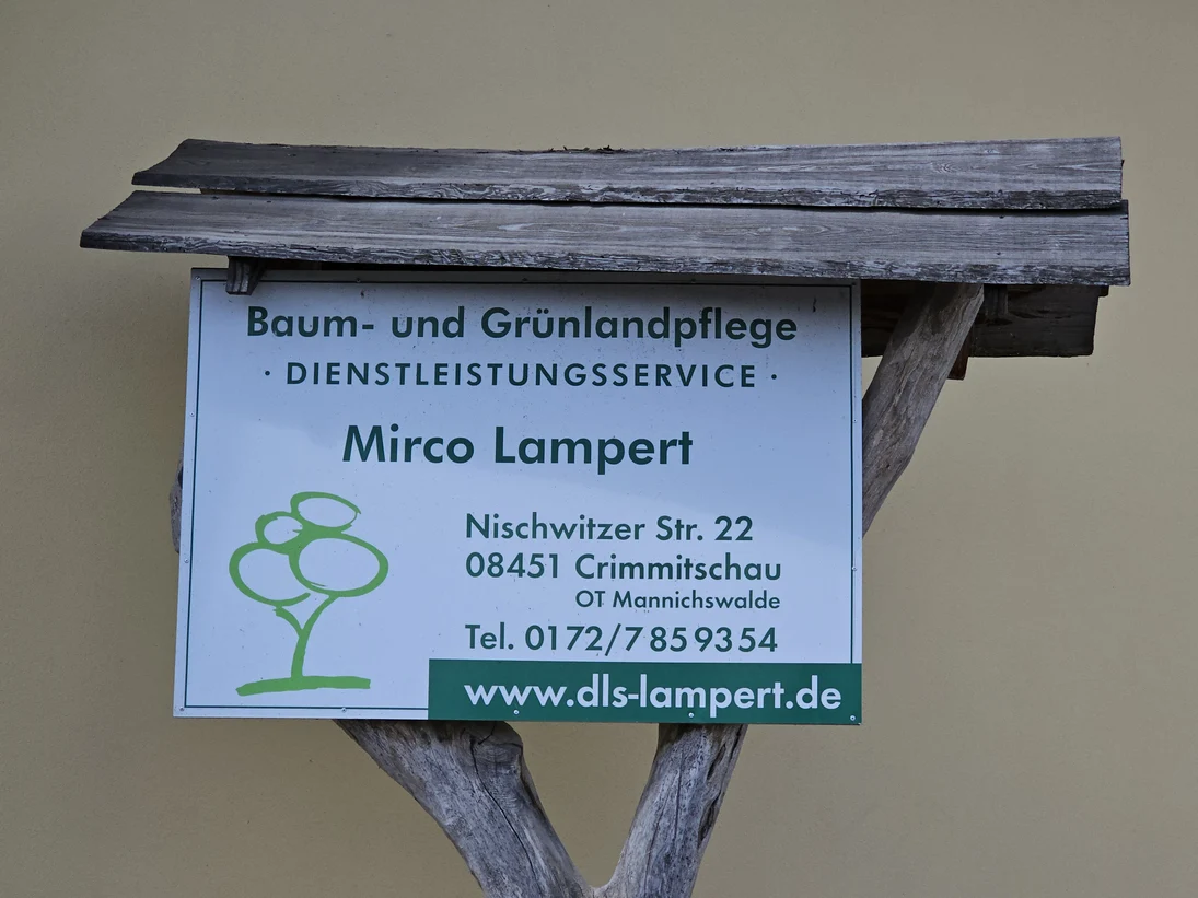 An einer Astgabel hängt das Firmenschild mit den Kontaktinformationen, geschützt von einem kleinen Holzdach
