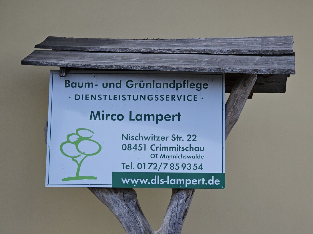 An einer Astgabel hängt das Firmenschild mit den Kontaktinformationen, geschützt von einem kleinen Holzdach