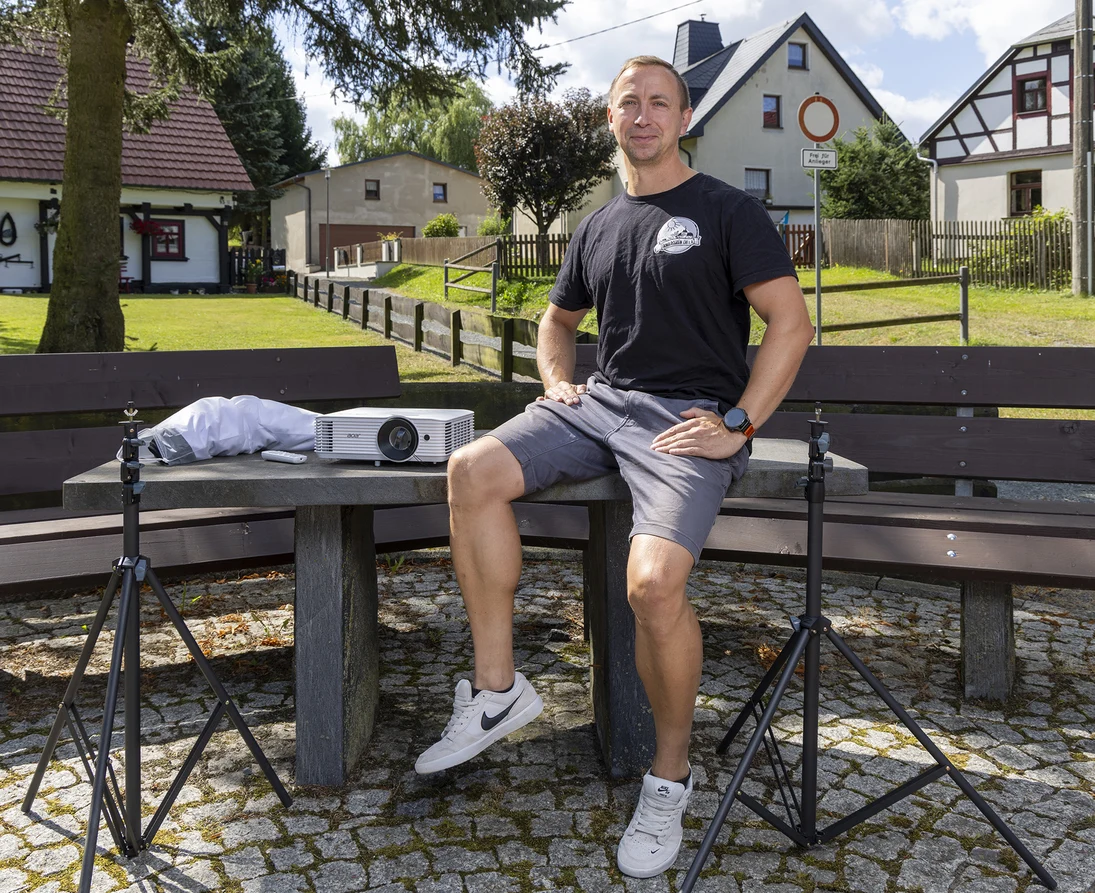 ein Mann sitzt auf einem Tisch auf dem Dorfplatz, neben sich zwei Stative und ein Beamer