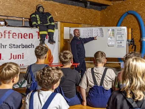 ein Feuerwehrmann steht an einer weißen Tafel und zeigt vor ihm sitzenden Kindern etwas