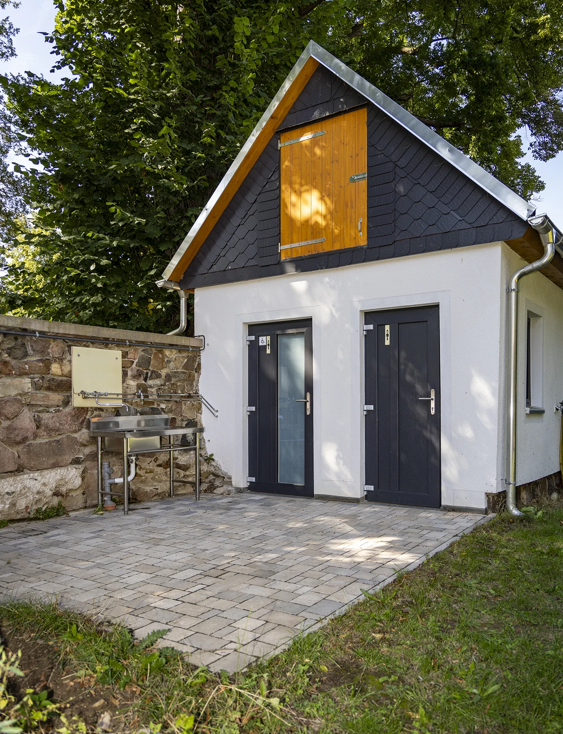 eine kleines Haus mit zwei Türen, im Hintergrund ist eine an einer Steinmauer angebrachte metallene Spüle zu sehen