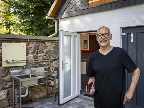 ein Mann steht vor einem kleinen Haus mit geöffneter Tür, im Hintergrund ist eine an einer Steinmauer angebrachte metallene Spüle zu sehen