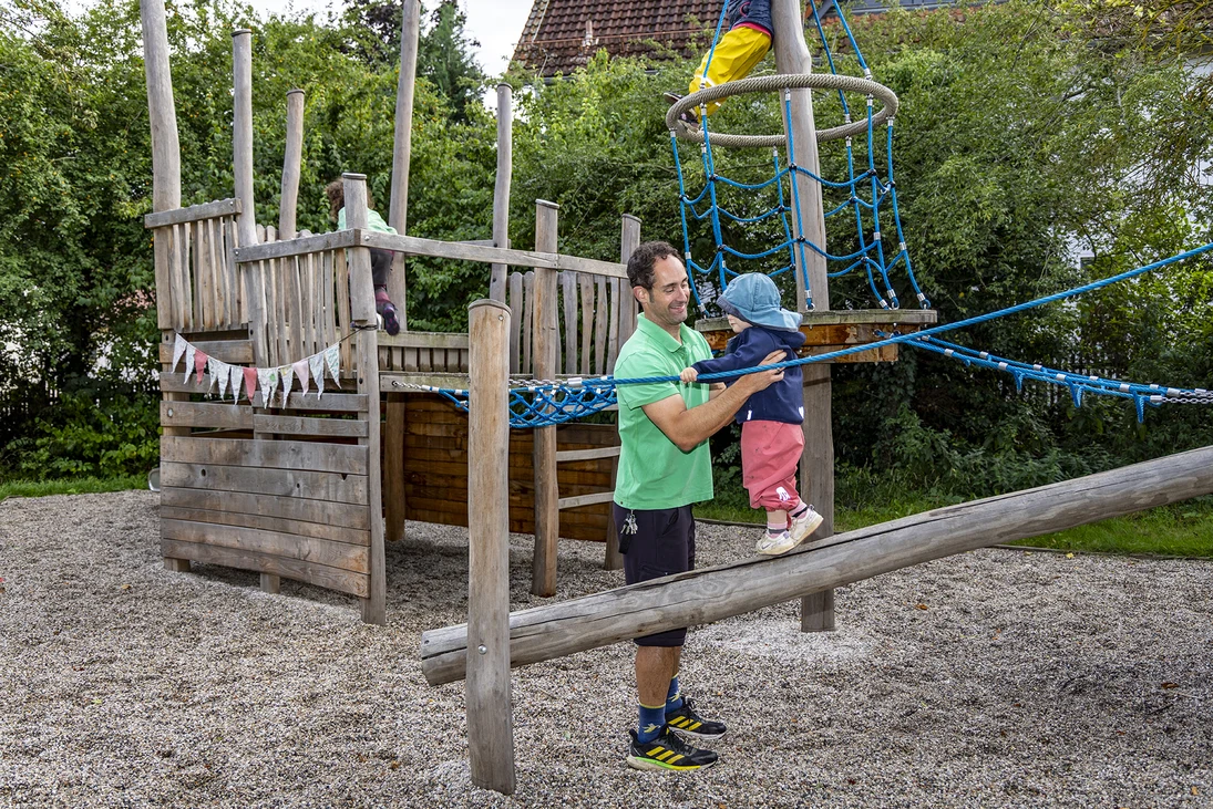 ein Mann hilft einem kleinen Kind beim Balancieren auf einem Holzbalken, im Hintergrund andere spielende Kinder auf einem großen hölzernen Spielschiff