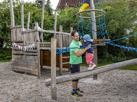ein Mann hilft einem kleinen Kind beim Balancieren auf einem Holzbalken, im Hintergrund andere spielende Kinder auf einem großen hölzernen Spielschiff