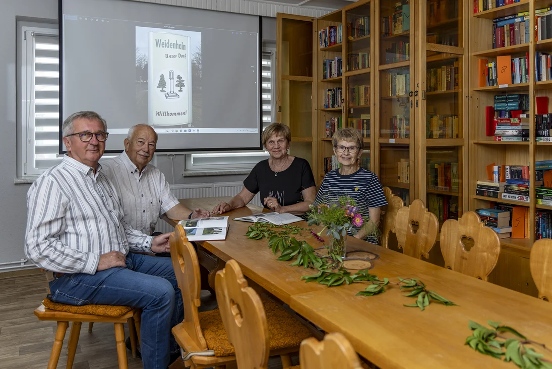 vier Menschen sitzen an einem Holztisch und haben geöffnete Bücher vor sich liegen, im Hintergrund ein gut gefüllter Bücherschrank