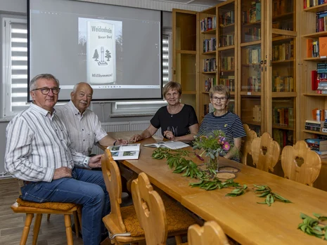 vier Menschen sitzen an einem Holztisch und haben geöffnete Bücher vor sich liegen, im Hintergrund ein gut gefüllter Bücherschrank