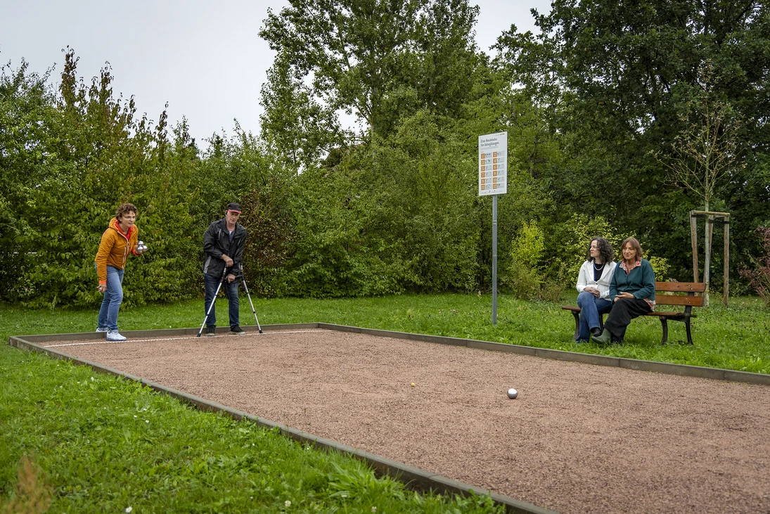 zwei Menschen sitzen am Ende einer Boule-Bahn, zwei sitzen auf einer Bank daneben, auf der Boule-Bahn liegt eine silberne Kugel