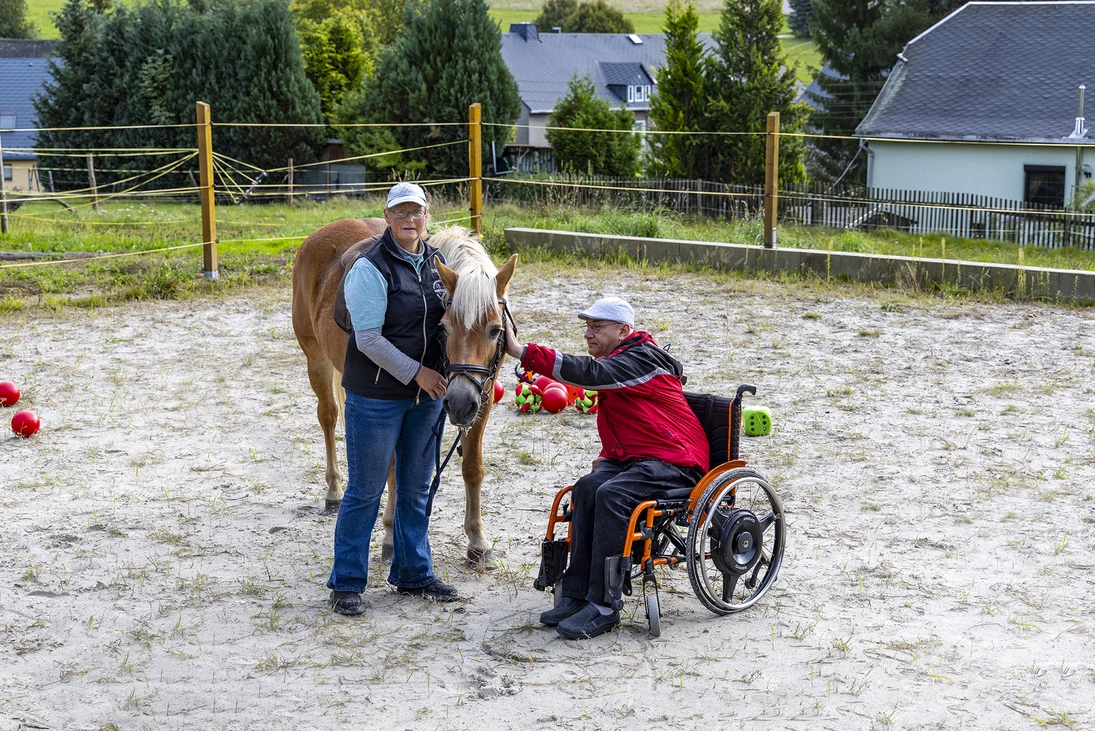 auf einem Reitplatz streichelt ein Rollstuhlfahrer ein Pferd, eine Frau steht daneben und hält das Pferd am Halfter