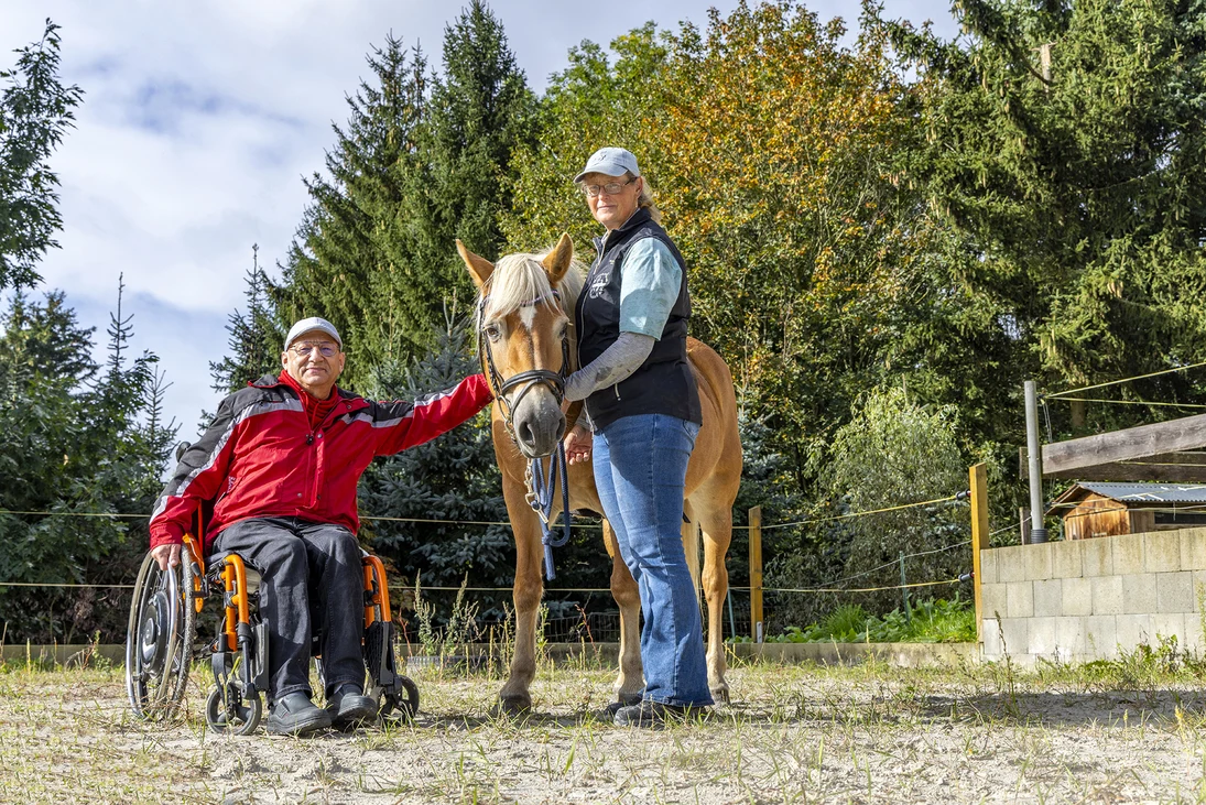 ein Rollstuhlfahrer streichelt ein Pferd, eine Frau steht daneben und hält das Pferd am Halfter
