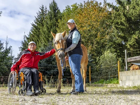 ein Rollstuhlfahrer streichelt ein Pferd, eine Frau steht daneben und hält das Pferd am Halfter