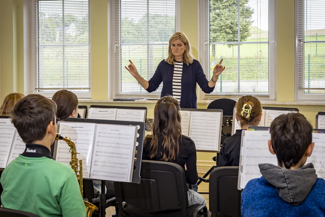 eine Dirigentin steht vor einem aus Kindern bestehenden Orchester, man sieht Blasinstrumente und Notenständer