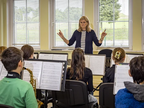 eine Dirigentin steht vor einem aus Kindern bestehenden Orchester, man sieht Blasinstrumente und Notenständer