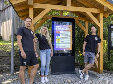 drei Menschen stehen vor einem offenem Holzbalkenbau mit Dach, unter dem eine elektronische Anzeigetafel aufgebaut ist