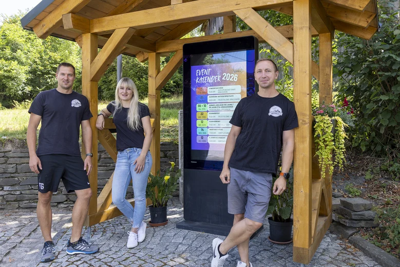 Auf dem Bild stehen drei Personen neben einer hölzernen Konstruktion, in der ein digitaler Bildschirm mit einem Veranstaltungskalender steckt.