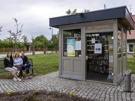 auf einer gepflasterten Fläche steht ein Bücherkiosk, links davon sitzen drei Frauen auf einer Metallbank