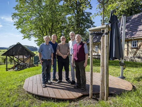 fünf Menschen stehen auf einer hölzernen Plattform auf einer Wiese, davor sieht man einen frisch gepflanzten Baum