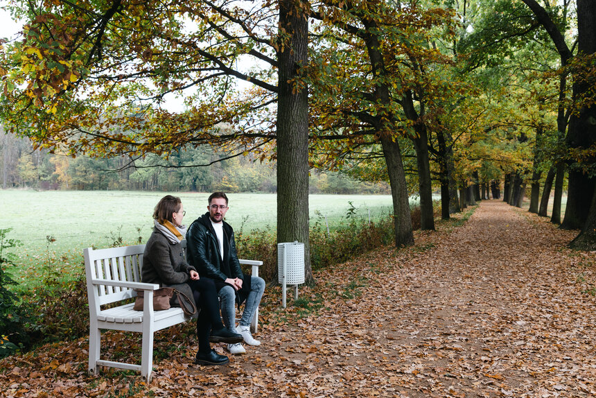 Eine Frau und ein Mann sitzen auf einer neuen Parkbank an einer Allee im Grünfelder Park 