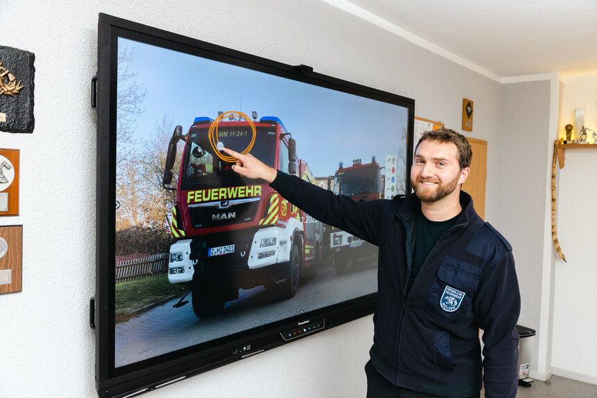 Ein Mitglied des Feuerwehrvereins deutet mit dem Finger stolz auf das neue interaktive Whiteboard, auf dem ein Feuerwehrauto gezeigt wird