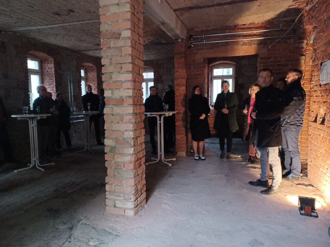 Einige Personen stehen im Keller des Gebäudes. In der Mitte ist eine Säule. An den Wänden und an der Säule sieht man die Ziegel.