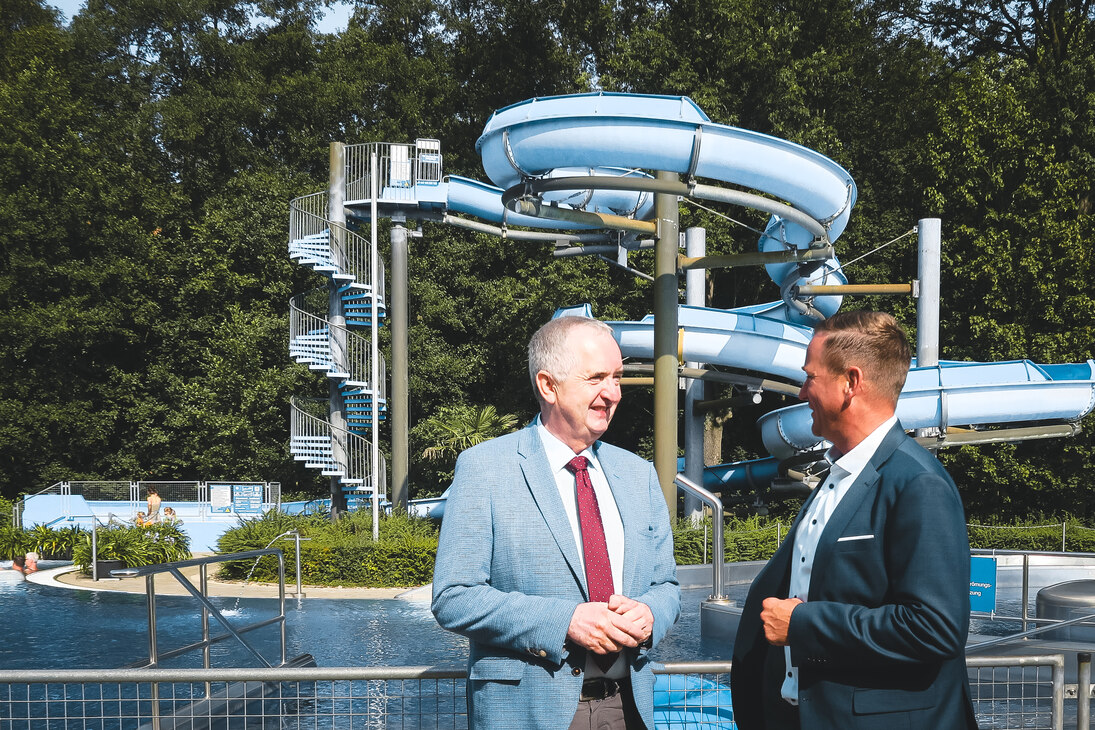 Staatsminister Schmidt und Bürgermeister Peters stehen im Freibad. Im Hintergrund ist ein Schwimmbecken sowie eine blaue Riesenrutsche zu sehen