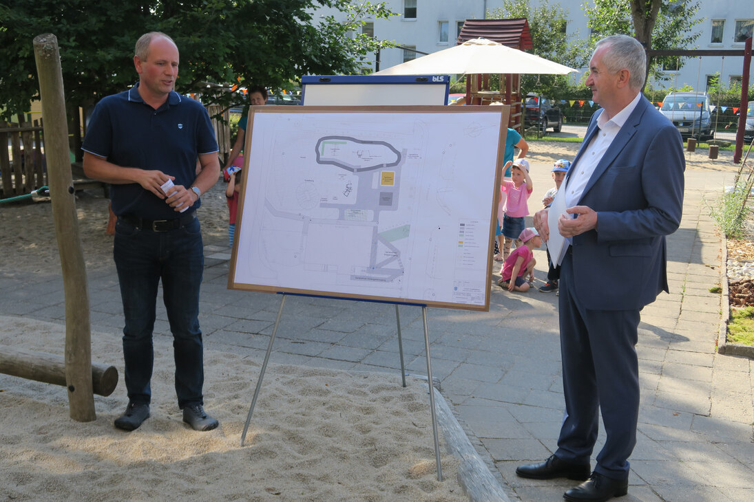 Denny Hantsche erläutert Staatsminister Schmidt die Neugestaltung der Außenanlagen. Beide stehen im Kita-Gelände vor einer Tafel mit der geplanten Maßnahme.