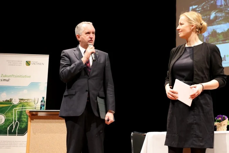 Auf dem Podium. Staatsminister Thomas Schmidt spricht in ein Mikrophon und richtet sein Wort an die rechts von ihm stehende Moderatorin Carmen Hentschel.