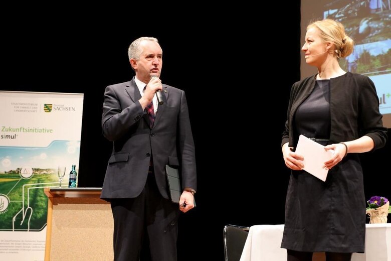 Auf dem Podium. Staatsminister Thomas Schmidt spricht in ein Mikrophon und richtet sein Wort an die rechts von ihm stehende Moderatorin Carmen Hentschel.