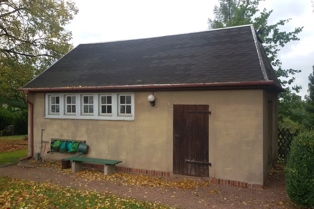 Unsanierte Trauerhalle in der Seitenansicht mit alter Holztür. Vor der Trauerhalle steht eine grüne Bank, 5 Gießkannen hängen an der Wand.