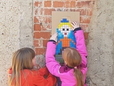 bunte Mosaikfiguren an einer Mauer. Davor zwei Mädchen.