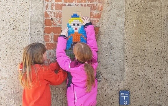 zwei Mädchen festigen ihr gestaltetes Mosaikbild an einer Ziegelwand