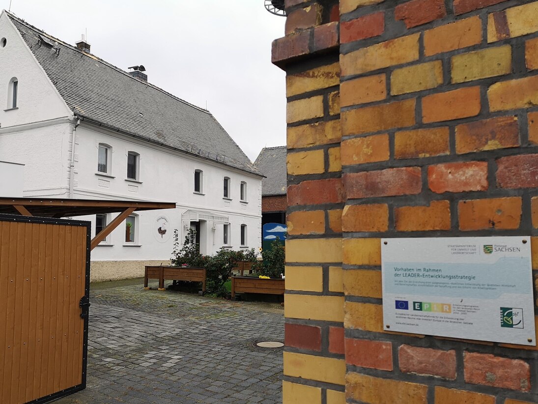 Blick durch das offene Hoftor auf das zweietagige Wohnhaus, rechts vom Tor hängt an der Wand eine Info-Tafel zur LEADER-Förderung