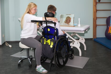 Eine Therapeutin sitzt auf einem Stuhl, neben ihr ein junger Mann im Rollstuhl. Er wird gerade therapeuitsch behandelt.