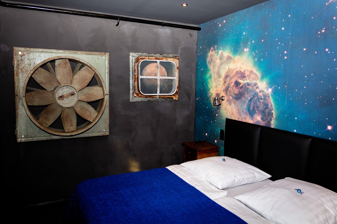 Raum mit Doppelbett, schwarzer Kopfteil, dunkelblaue Tagesdecke. Wand im Kopfteil des Bettes: Weltalldarstellung, Sterne, ein großer Meteorit. Decke und Wand hinterm Bett schwarz. An dieser Ventilator-Propeller und Fenster mit "Blick" auf einen Planeten.