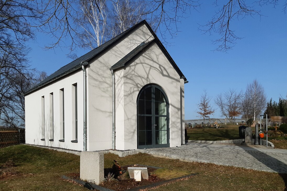 Haupteingang der Trauerhalle, umgeben von Birkenbäumen. Weiße Wände, schmale hohe Fenster an den Seitenwänden, Vorbau an der Giebelseite mit einer hohen verglasten abgerundeten Tür.