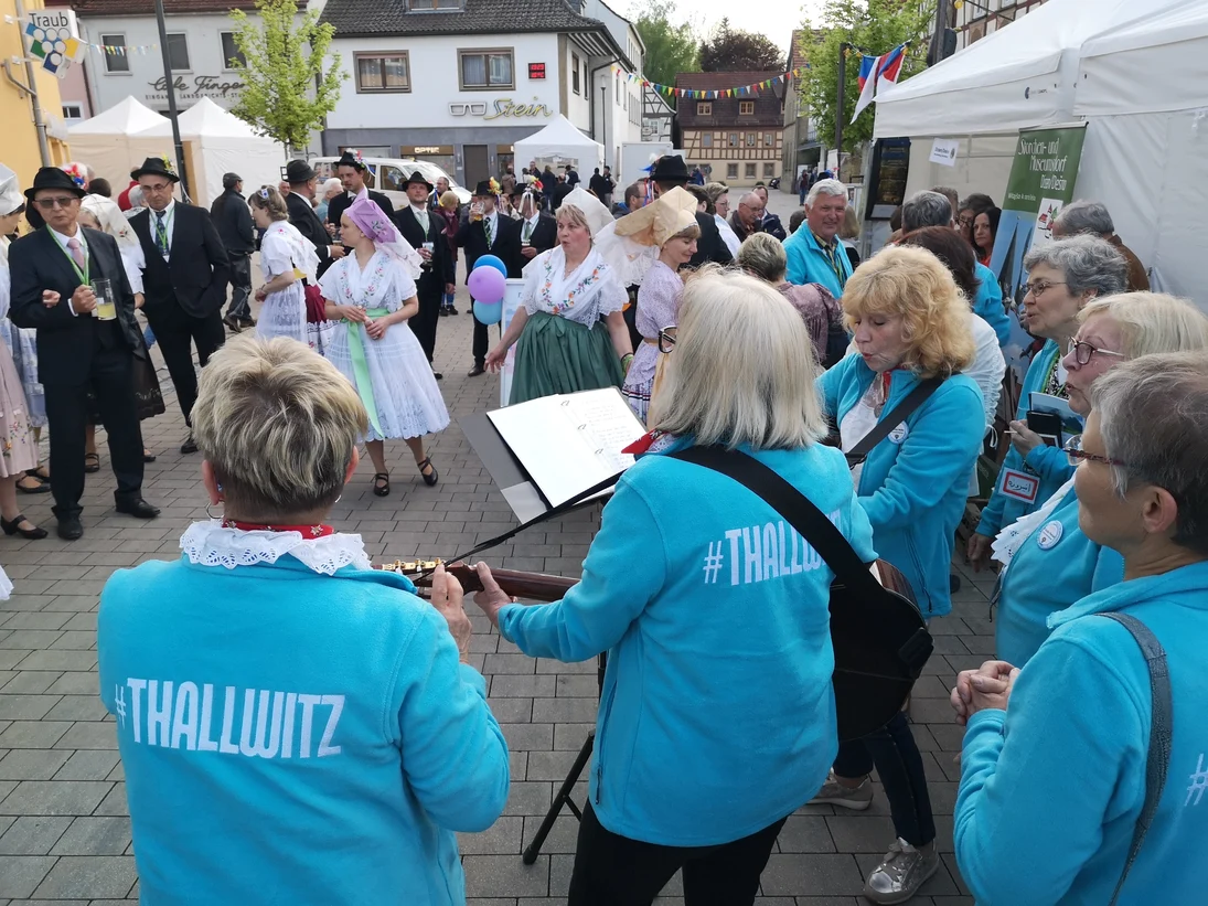 Frauen in hellblauen Jacken mit Aufdruck #Thallwitz spielen Gitarre und singen vor Publikum auf einer Straße