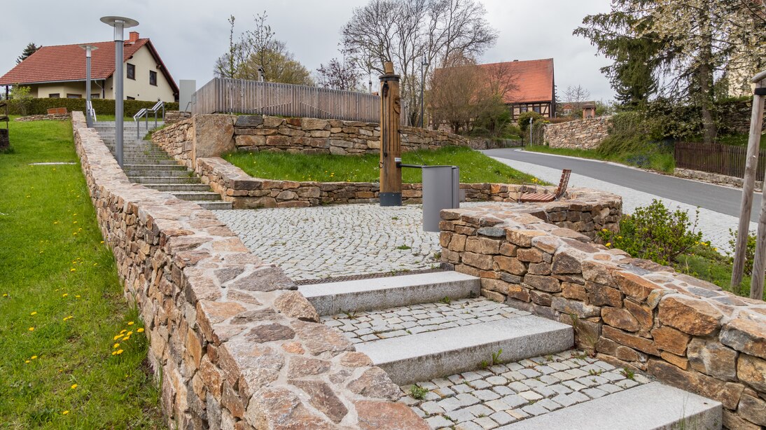 Eine Treppe überbrück die Hanglage, langgezogene Podeststufen, Granitstufen und -pflasterung, durch eine gemauerte Feldsteinmauer gesäumt